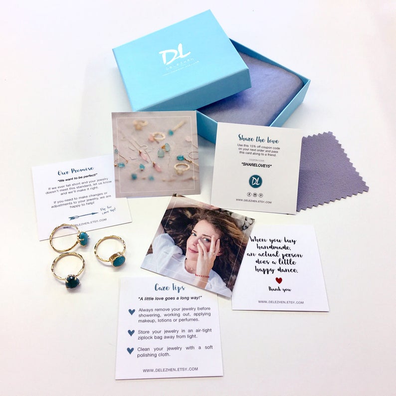 May include: A set of three turquoise and gold rings in a small white box with a blue lid. The box is surrounded by several small cards with text about the jewelry and the shop. One card says "Our Promise" and another card says "Care Tips".