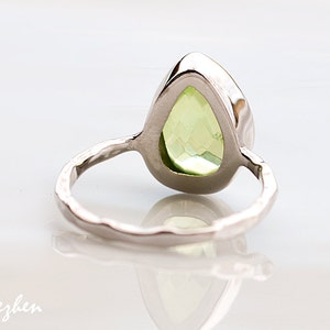 Pu&ograve; includere: Un anello d'argento con una pietra preziosa verde chiaro a forma di pera. L'anello ha una fascia strutturata e un'incastonatura lucida. L'anello &egrave; esposto su una superficie bianca. Gioielleria.
