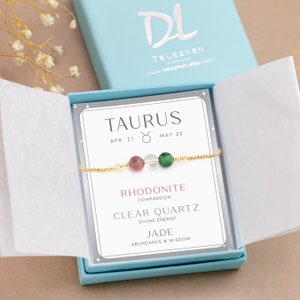 May include: A light blue gift box with a white card inside. The card has the zodiac sign Taurus with the dates April 21 to May 20. The card also lists the stones in the bracelet: Rhodonite, Clear Quartz, and Jade, with their corresponding meanings. The bracelet is made of gold chain with three beads: pink, clear, and green.