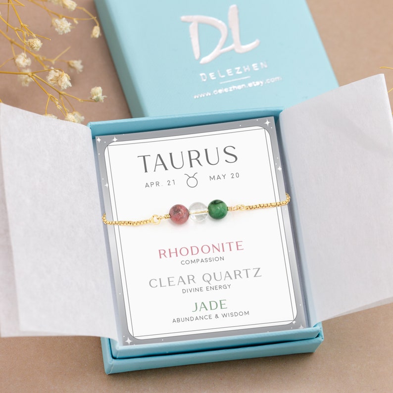 May include: A light blue gift box with a white card inside. The card has the zodiac sign Taurus with the dates April 21 to May 20. The card also lists the stones in the bracelet: Rhodonite, Clear Quartz, and Jade, with their corresponding meanings. The bracelet is made of gold chain with three beads: pink, clear, and green.