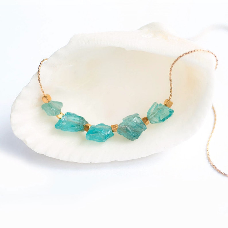May include: A gold chain necklace with turquoise blue gemstone beads. The necklace is displayed on a white seashell.