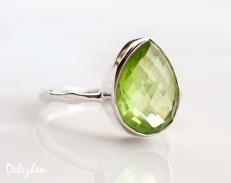 Pu&ograve; includere: Un anello d'argento con una pietra preziosa verde chiaro a forma di pera e sfaccettata. La pietra &egrave; incastonata in una fascia d'argento liscia e lucida. L'anello &egrave; esposto su una superficie bianca.