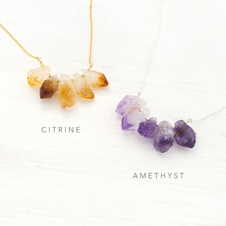 May include: Two necklaces with raw crystal pendants. The top necklace features a cluster of six citrine stones in shades of yellow and orange. The bottom necklace features a cluster of five amethyst stones in shades of purple. The text "CITRINE" is below the top necklace and the text "AMETHYST" is below the bottom necklace.