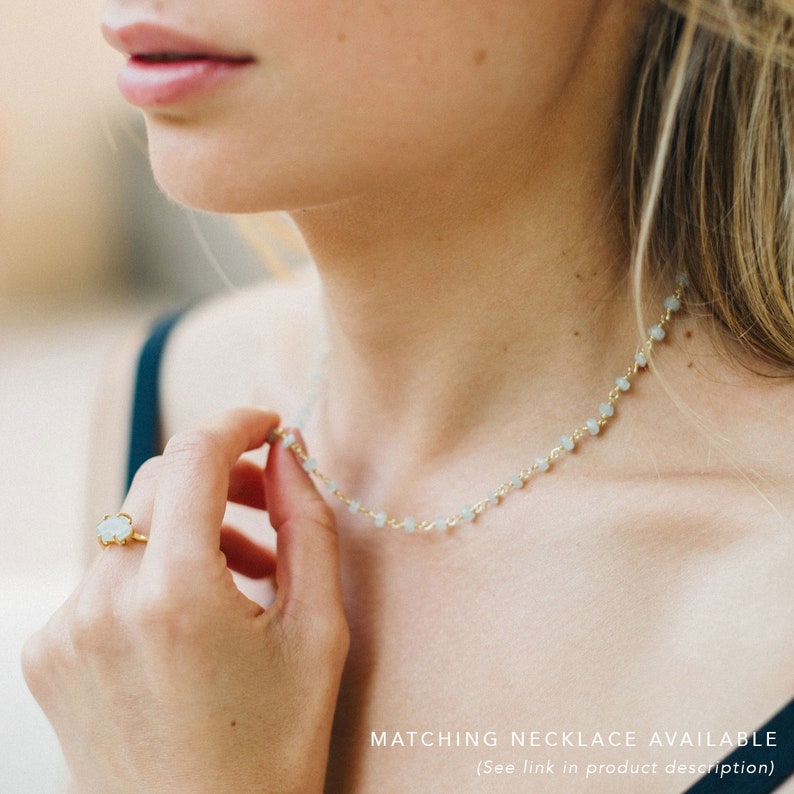 K&ouml;nnte beinhalten: Gezeigt wird eine zarte goldene Halskette mit kleinen, hellblauen Perlen. Ein passender Ring mit einem hellblauen Stein ist ebenfalls zu sehen. Der Text "MATCHING NECKLACE AVAILABLE" befindet sich am unteren Bildrand.