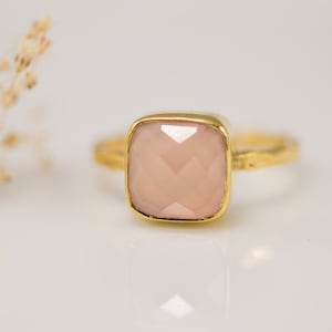 May include: A gold ring featuring a square, pale pink gemstone. The ring is displayed against a white backdrop.