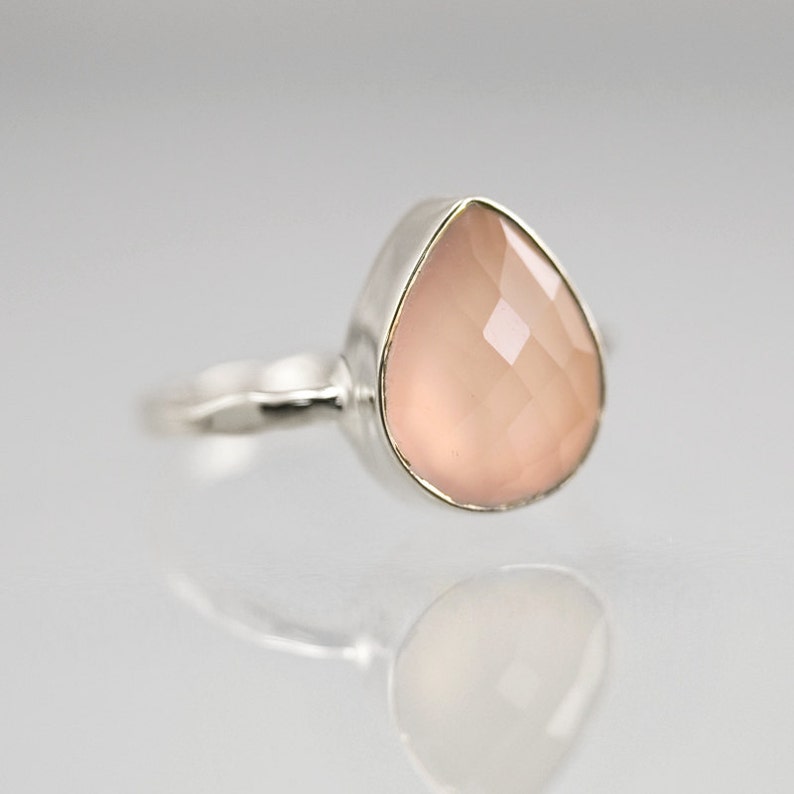 K&ouml;nnte beinhalten: Ein silberner Ring mit einem tropfenf&ouml;rmigen, facettierten, hellrosa Edelstein. Das Band des Rings hat ein strukturiertes Design. Der Edelstein ist in einer silbernen L&uuml;nette gefasst.