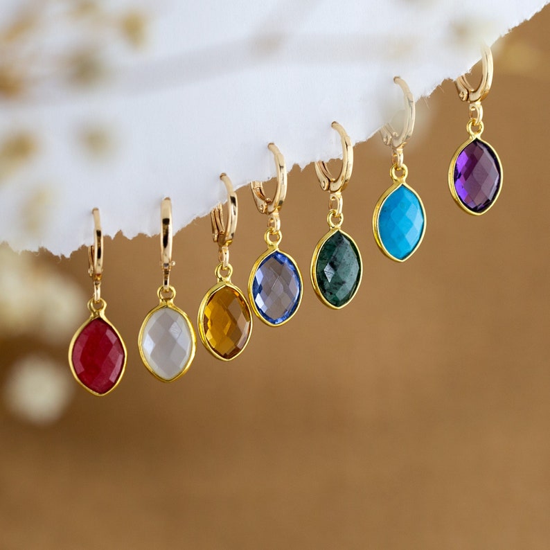May include: A set of eight gold hoop earrings with dangling gemstone charms. The charms are in various colors, including red, white, yellow, blue, green, turquoise, and purple.