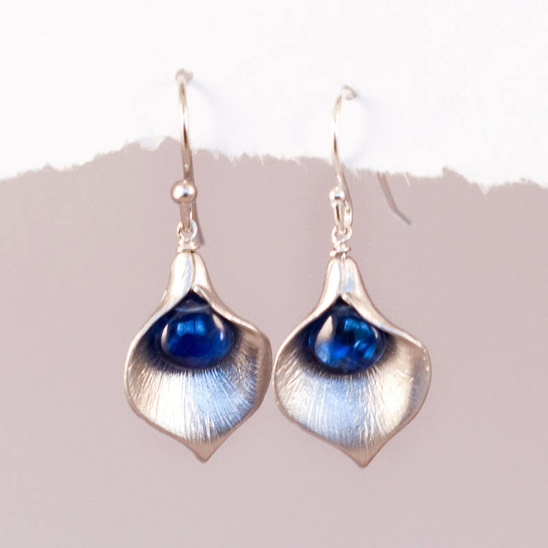 May include: A pair of silver earrings with a blue gemstone in the center of each earring. The earrings are shaped like a flower petal.