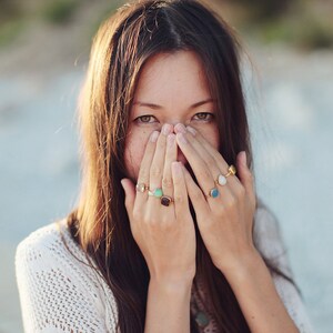 May include: A woman with long brown hair is covering her face with her hands. She is wearing several rings with colorful gemstones on her fingers.
