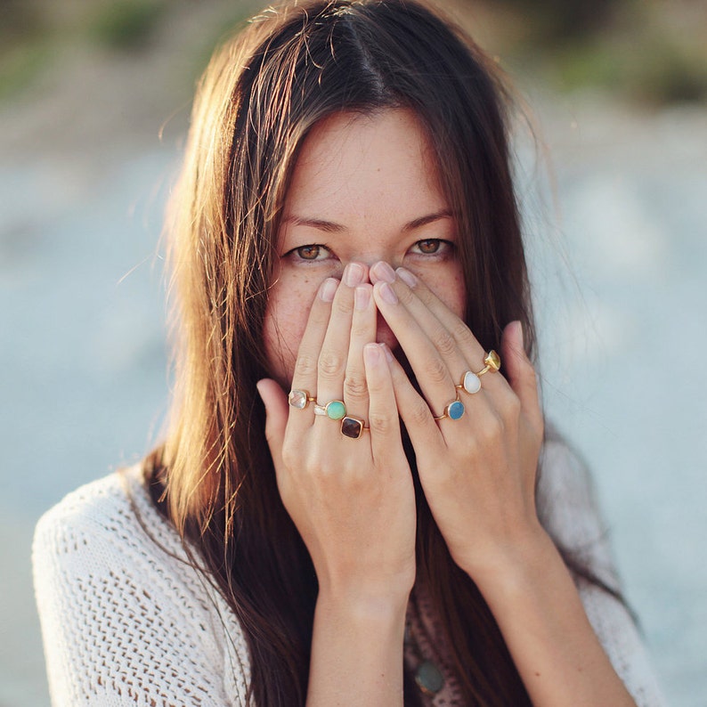 May include: A woman with long brown hair is covering her face with her hands. She is wearing several rings with colorful gemstones on her fingers.