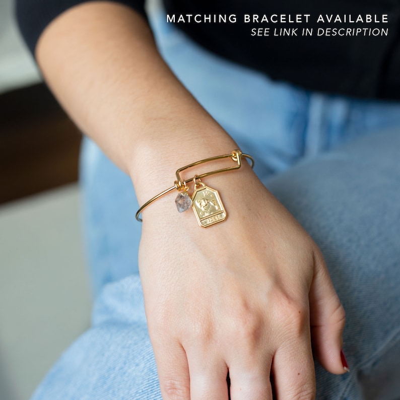 May include: Gold bracelet with a charm featuring the zodiac sign Gemini and a small clear crystal. The bracelet is on a person's wrist.