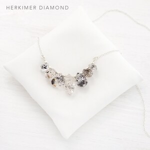 May include: A silver necklace with a cluster of clear quartz crystals, known as Herkimer diamonds, hanging from the chain. The necklace is displayed on a white pillow.