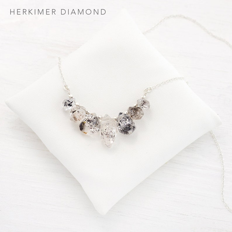 May include: A silver necklace with a cluster of clear quartz crystals, known as Herkimer diamonds, hanging from the chain. The necklace is displayed on a white pillow.