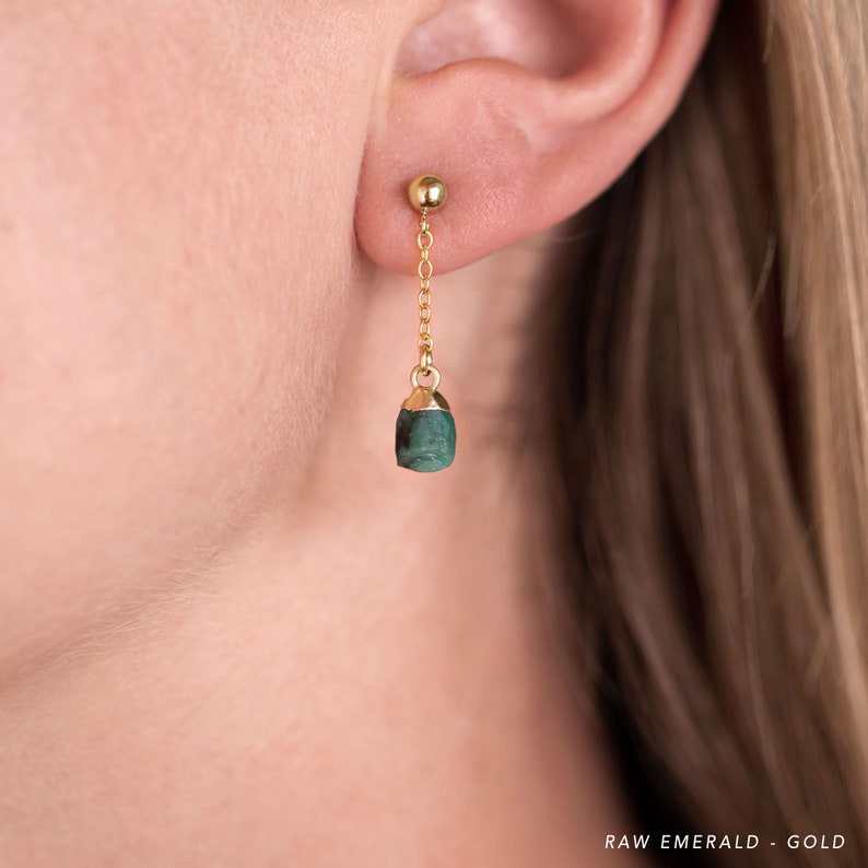 以下が含まれることがあります： 繊細なチェーンからぶら下がった、生のエメラルドストーンが付いたゴールドのイヤリング。エメラルドは深い緑色で、自然な研磨されていない外観をしています。