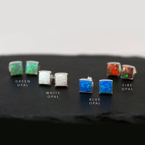 Pu&ograve; includere: Sei paia di orecchini a perno d'argento con pietre di opale quadrate in vari colori: verde, bianco, blu e arancione. Gli orecchini sono esposti su una superficie nera con i nomi dei colori scritti sotto ogni coppia.