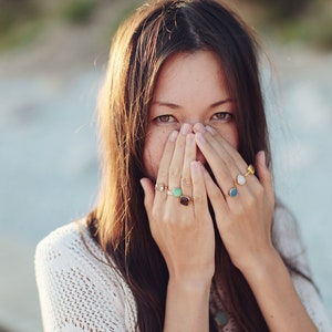 Puede incluir: Una mujer con el pelo largo y casta&ntilde;o se tapa la boca con las manos. Lleva varios anillos en los dedos, incluyendo un anillo con una piedra verde, un anillo con una piedra azul y un anillo con una piedra blanca.