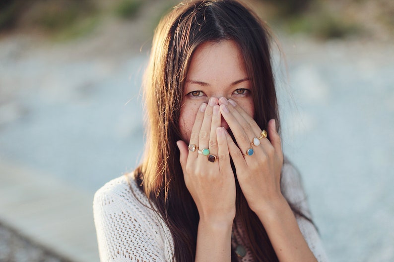 Puede incluir: Una mujer con el pelo largo y casta&ntilde;o se tapa la boca con las manos. Lleva varios anillos en los dedos, incluyendo un anillo con una piedra verde, un anillo con una piedra azul y un anillo con una piedra blanca.