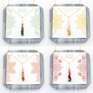 May include: Four gold necklaces with different colored stones, each on a white card with a mandala design and the name of a chakra. The necklaces are labeled "Heart Chakra", "Solar Plexus Chakra", "Sacral Chakra", and "Root Chakra".