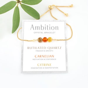 May include: A gold-colored adjustable bracelet with three gemstone beads: brown rutilated quartz, orange carnelian, and yellow citrine. The card behind the bracelet reads "Ambition Crystal Bracelet" and lists the properties of each stone.