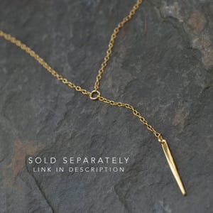 Op de afbeelding: Een gouden ketting met een lange, dunne, puntige hanger. De ketting ligt op een grijs, getextureerd oppervlak. APART VERKOCHT LINK IN DE BESCHRIJVING