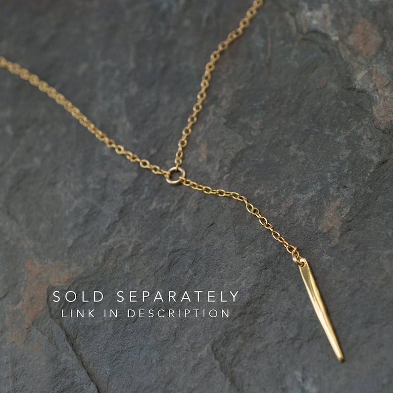 Op de afbeelding: Een gouden ketting met een lange, dunne, puntige hanger. De ketting ligt op een grijs, getextureerd oppervlak. APART VERKOCHT LINK IN DE BESCHRIJVING