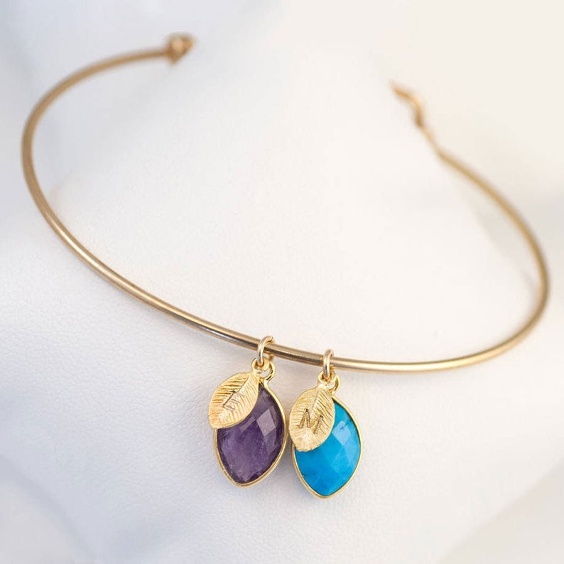 May include: Gold bangle bracelet with two charms. Each charm features a gold leaf with an initial and a gemstone. One charm has a purple amethyst and the other has a turquoise gemstone.