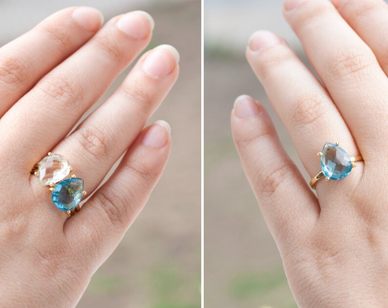 May include: Two teardrop-shaped rings with gold settings. The ring on the left has a clear stone, and the ring on the right has a light blue stone.