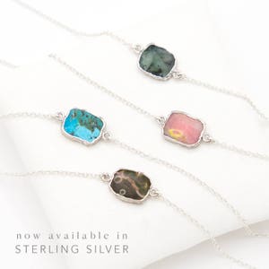 May include: Four silver chain necklaces with different colored stone pendants. The stones are turquoise, pink, green, and brown. The necklaces are displayed on a white surface. "now available in STERLING SILVER" is written below the necklaces.