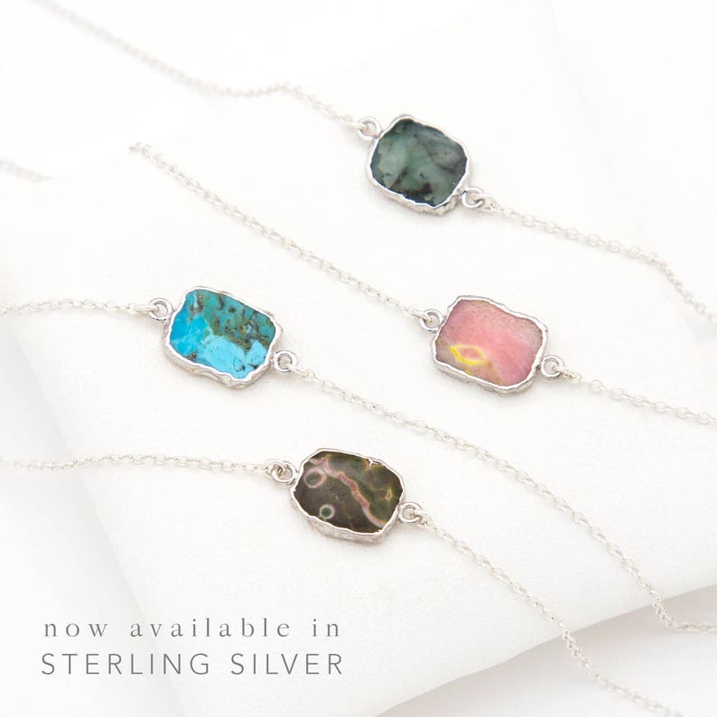 May include: Four silver chain necklaces with different colored stone pendants. The stones are turquoise, pink, green, and brown. The necklaces are displayed on a white surface. "now available in STERLING SILVER" is written below the necklaces.