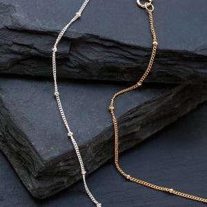 Puede incluir: Dos delicados collares con pequeñas cuentas, uno plateado y otro dorado, sobre un fondo gris oscuro.