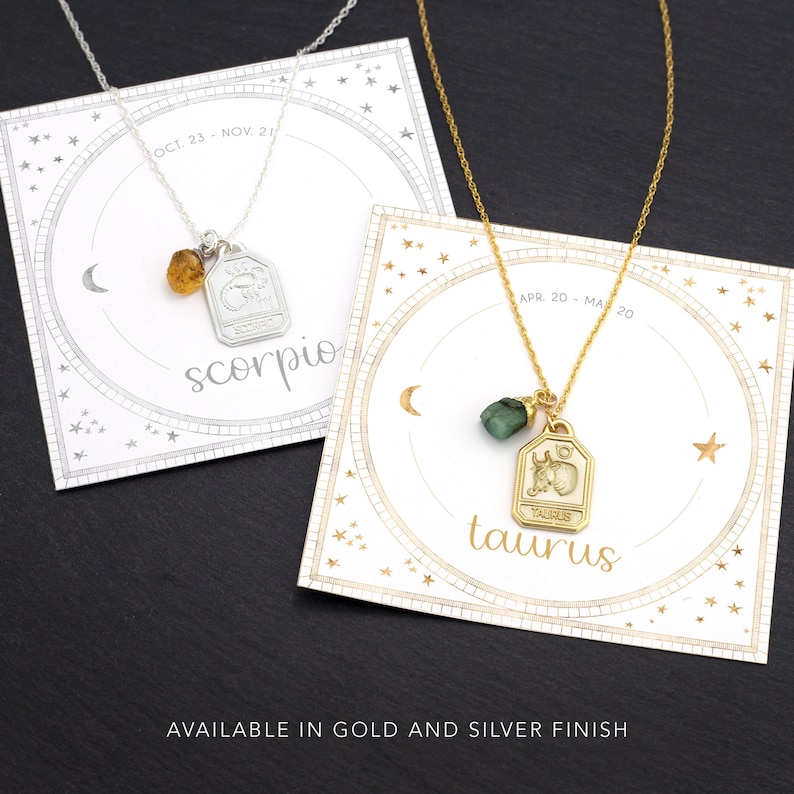 May include: Two gold necklaces with zodiac signs, Scorpio and Taurus, on white cards with gold stars. The Scorpio necklace has an amber stone and the Taurus necklace has a green stone.