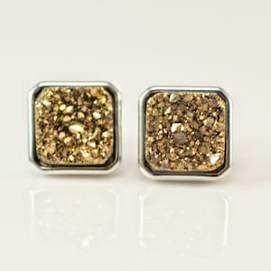 May include: A pair of gold and silver square stud earrings with a sparkly, textured surface.