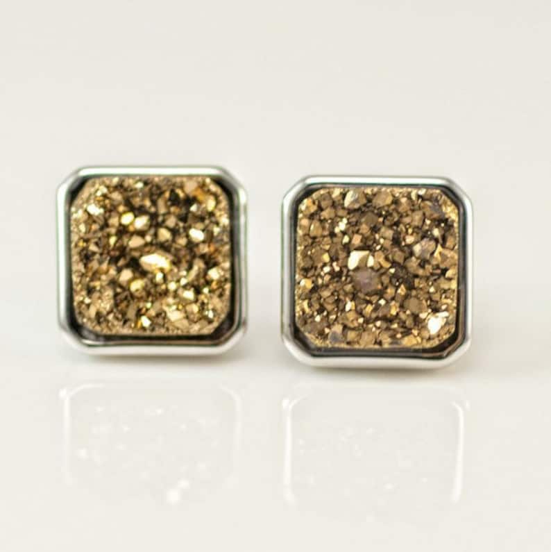 May include: A pair of gold and silver square stud earrings with a sparkly, textured surface.