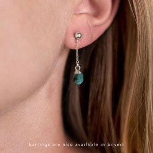 以下が含まれることがあります： 小さな緑色の宝石がチェーンからぶら下がっている、銀製のイヤリングのペア。イヤリングは人の耳につけられています。