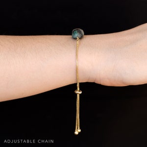 May include: A gold chain bracelet with a dark green gemstone bead. The bracelet has an adjustable chain with a sliding bead.