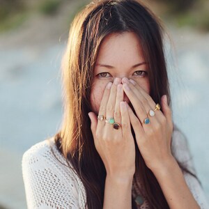 May include: A woman wearing a white sweater with a variety of colorful rings on her fingers. The rings are made of gold and have different shaped stones.