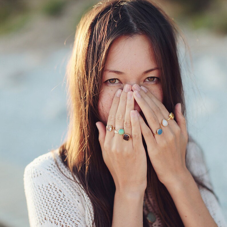 May include: A woman wearing a white sweater with a variety of colorful rings on her fingers. The rings are made of gold and have different shaped stones.