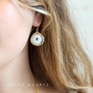 May include: Gold earrings with a white and gray solar quartz stone. The stone has a natural, crystal-like appearance.  The earrings are a teardrop shape and have a simple, elegant design.  "SOLAR QUARTZ" is written in the image.