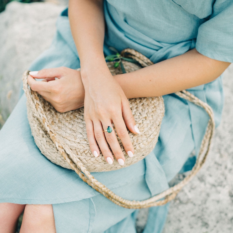 May include: A woman wearing a light blue dress with a woven straw bag. She is wearing a ring with a large green gemstone.
