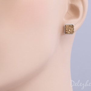 May include: A pair of silver stud earrings with a square-shaped gold glitter design.