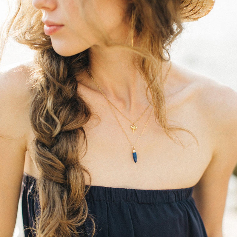 May include: A woman wearing a gold necklace with a small bird pendant and a larger blue gemstone pendant.