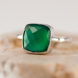 May include: A silver ring with a square green gemstone. The ring is set on a light brown surface.