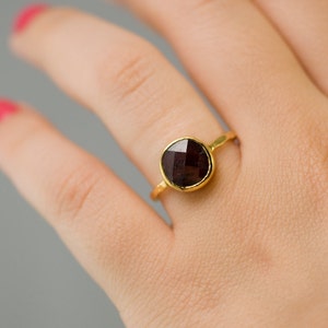 May include: A gold ring with a dark red gemstone. The ring is on a person's finger.