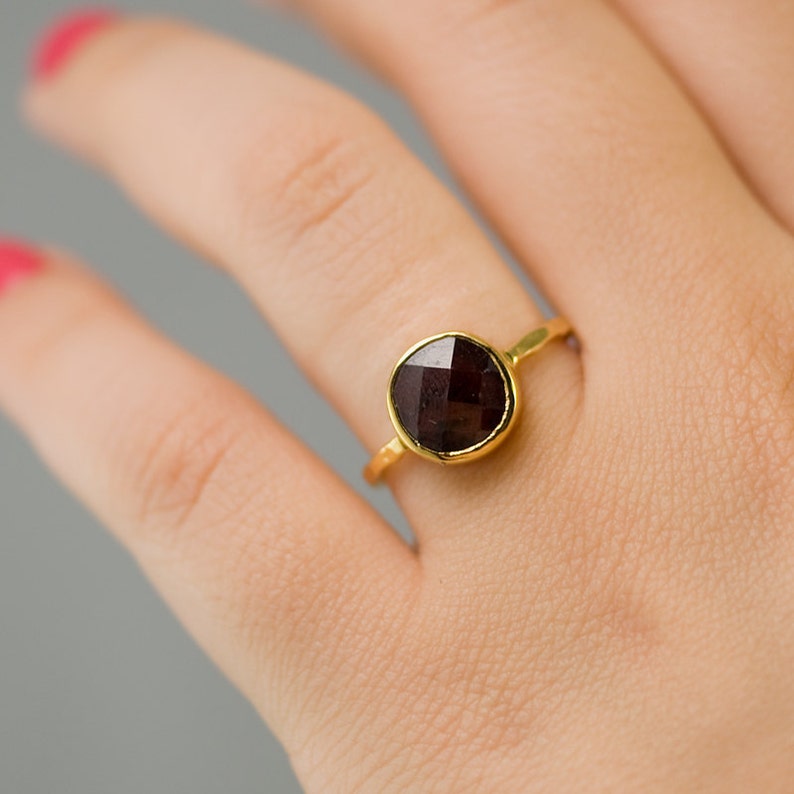 May include: A gold ring with a dark red gemstone. The ring is on a person's finger.