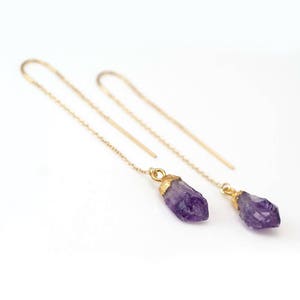 Puede incluir: Un par de pendientes de cadena dorada con gotas de cristal de amatista. Los cristales son en bruto y tienen una forma natural irregular.