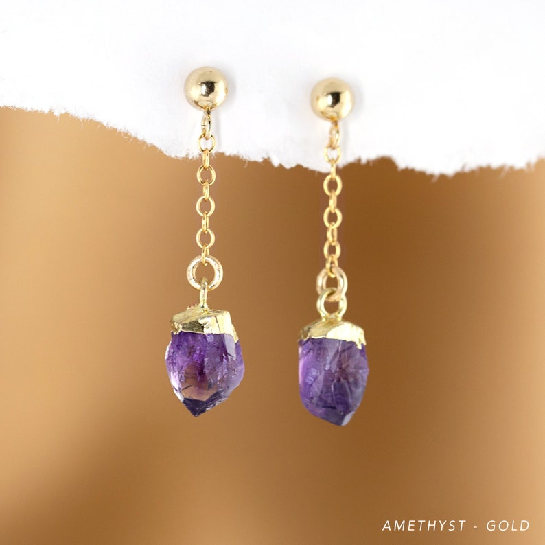 以下が含まれることがあります： ゴールドのイヤリングで、それぞれにアメジストのクリスタルがぶら下がっています。クリスタルは原石で、自然な不規則な形をしています。イヤリングはシンプルでエレガントです。