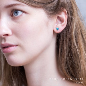 Puede incluir: Un par de pendientes de bot&oacute;n en forma de coraz&oacute;n de &oacute;palo azul verdoso. Los pendientes los lleva una persona con el pelo rubio.