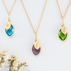May include: Three gold necklaces with leaf-shaped pendants. Each pendant has a coloured gemstone: blue, purple, and green. A small gold leaf with a letter is attached to each gemstone. The necklaces are on a white background.