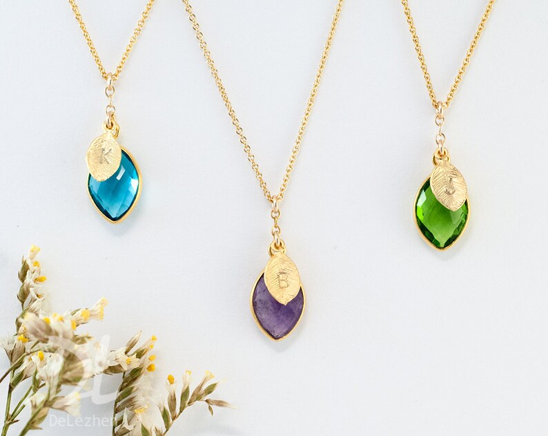 May include: Three gold necklaces with leaf-shaped pendants. Each pendant has a coloured gemstone: blue, purple, and green. A small gold leaf with a letter is attached to each gemstone. The necklaces are on a white background.