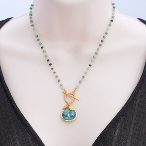 May include: A gold necklace with a green gemstone pendant and a toggle clasp. The necklace is made of small green beads and has a delicate, feminine look.